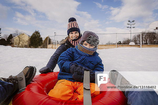 Portrait of mother and son tobogganing with tube sled on snow covered field against cloudy sky