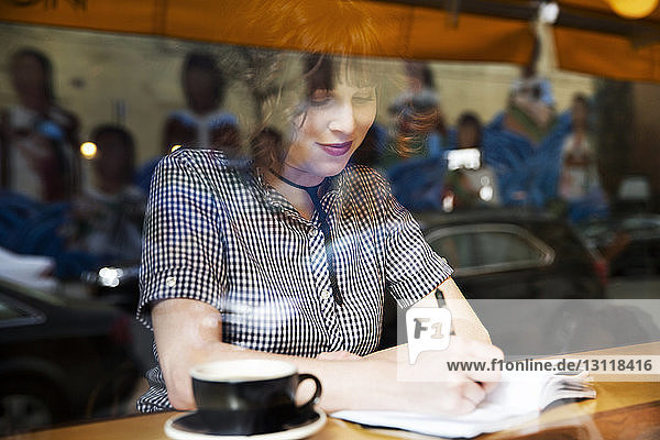 Frau schreibt auf Buch  während sie im Café sitzt und durch ein Fenster gesehen wird
