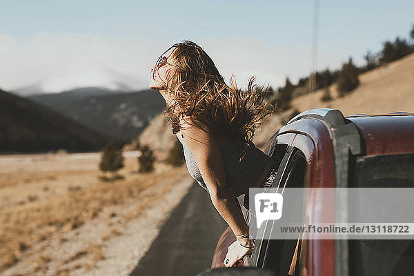 Seitenansicht einer sorglosen jungen Frau mit zerzaustem Haar  die sich aus dem Autofenster lehnt
