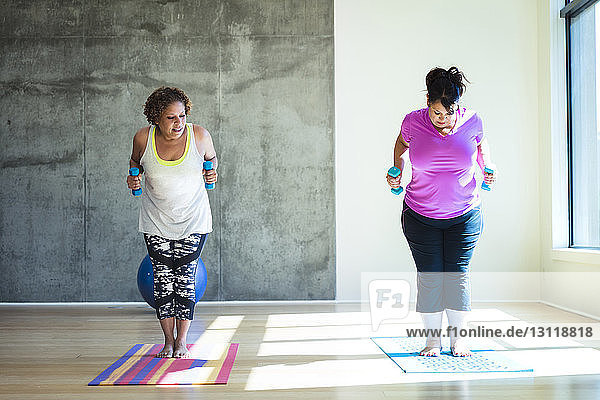 Woman looking at friend standing on exercise mat while holding dumbbells against wall in yoga studio