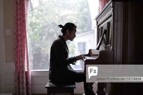 Seitenansicht eines Mannes  der zu Hause Klavier gegen ein Fenster spielt
