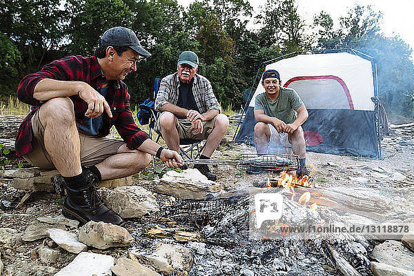 Mittelgroßer Mann kocht Fisch am Lagerfeuer  während er bei Sonnenuntergang mit Freunden zeltet