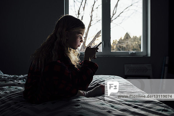 Seitenansicht eines Mädchens  das ein Mobiltelefon benutzt  während es zu Hause auf dem Bett sitzt