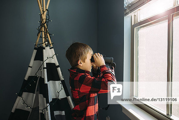 Seitenansicht eines Jungen  der zu Hause am Fenster durch ein Fernglas schaut