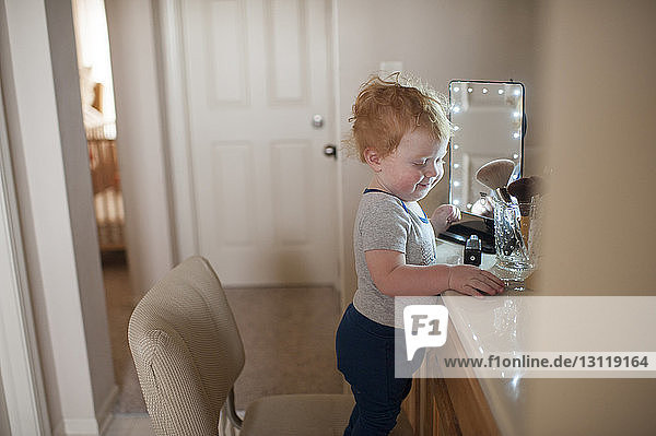 Side view of cute baby boy standing by beauty products on chair at home