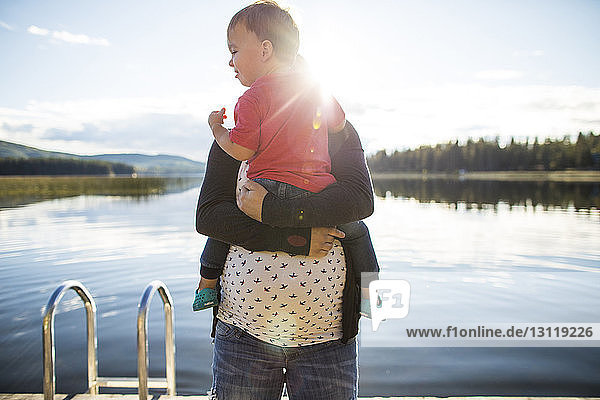 Pregnant mother carrying son by lake during sunny day