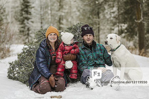 Porträt von Eltern mit Tochter und Hund an einer Kiefer sitzend auf einem schneebedeckten Feld im Wald