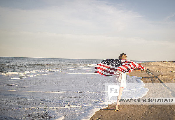 Rückansicht eines Teenagers  der die amerikanische Flagge hält  während er am Strand gegen den Himmel rennt