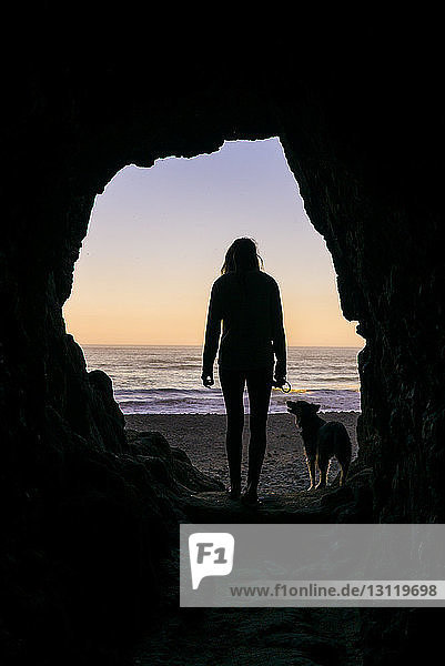 Silhouette woman with dog standing at cave entrance against beach