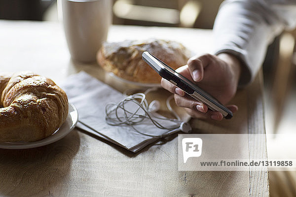 Cropped image of man holding mobile phone by croissants and coffee at table