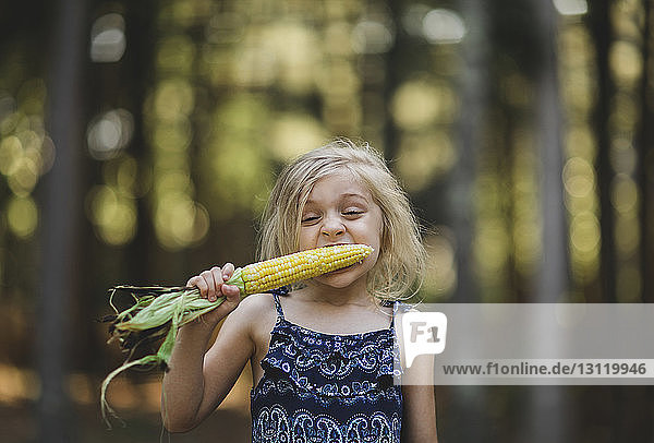Girl eating corn while standing against trees in yard