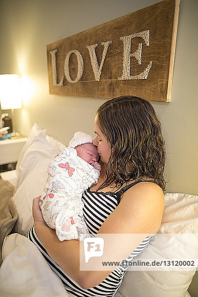 Mutter küsst neugeborene Tochter  während sie zu Hause im Bett sitzt