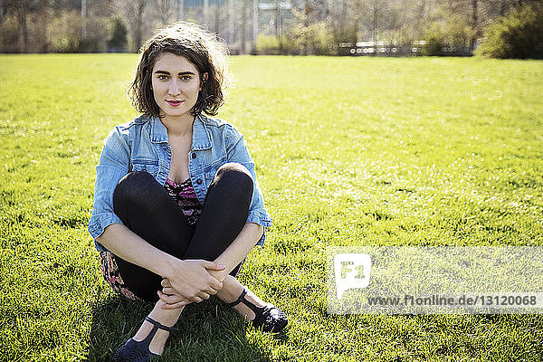 Portrait of confident woman hugging knees while sitting on grassy field