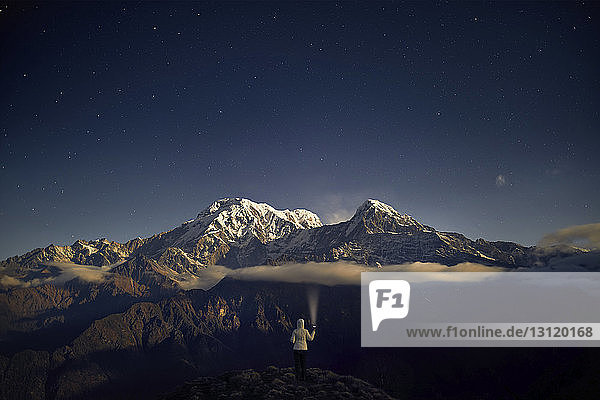 Rückansicht eines Touristen  der eine Taschenlampe hält  während er nachts auf einem Berg gegen den Himmel steht