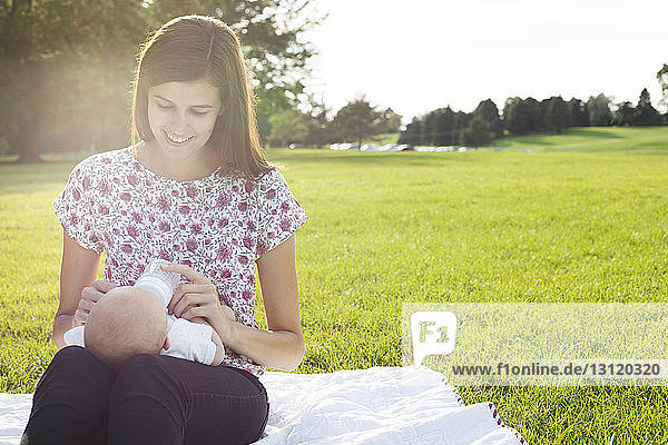 Lächelnde Mutter füttert Baby  während sie auf einem Grasfeld im Park sitzt