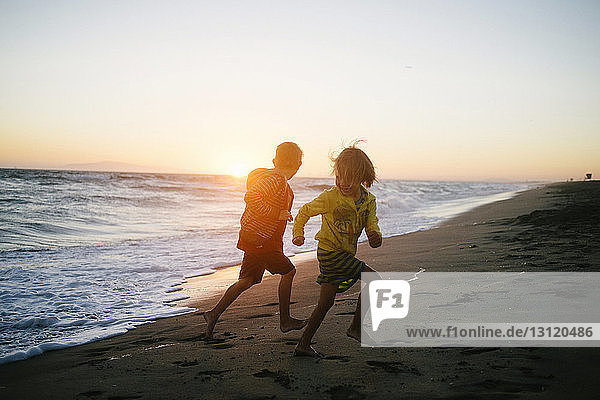 Siblings running on shore at beach against sky during sunset