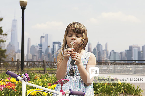 Cute girl blowing dandelion on promenade with city skyline in background