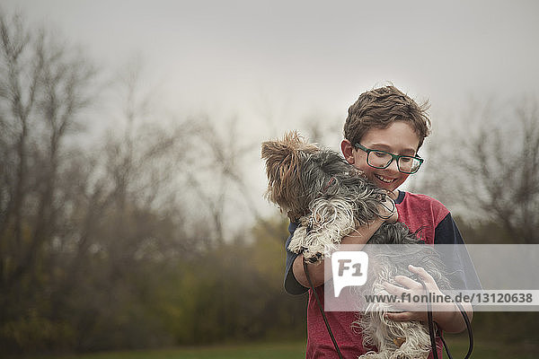 Glücklicher Junge trägt Hund  während er im Park gegen den klaren Himmel steht