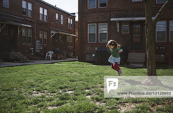 Verspieltes Mädchen in voller Länge  das auf Gras gegen ein Haus im Vorgarten springt