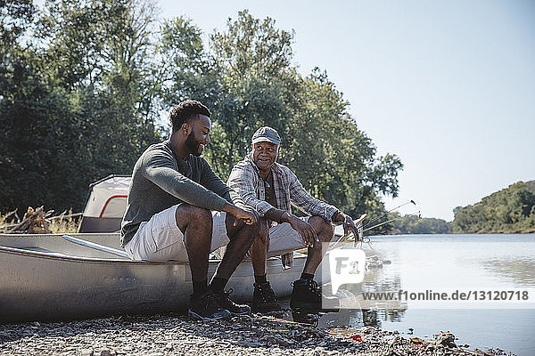 Male friends talking while sitting on boat at lakeshore against clear sky