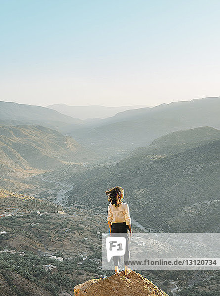 Rear view of woman looking at mountains while standing on rock against clear sky