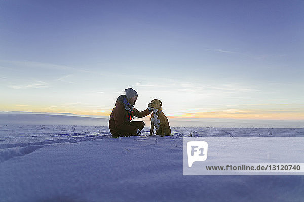 Hiker petting dog while sitting on snowy field against sky during sunset