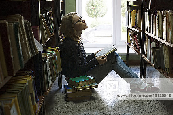 Frau hält Buch und schaut auf  während sie in der Bibliothek sitzt