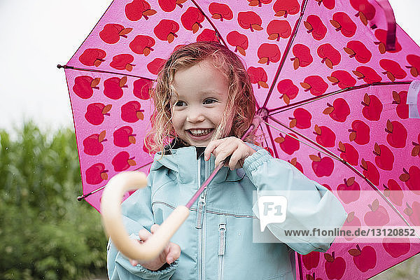 Glückliches niedliches Mädchen mit rosa Regenschirm  das während der Regenzeit im Freien steht