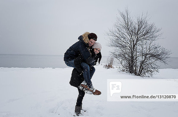Glückliche Frau nimmt einen männlichen Freund huckepack  während sie am schneebedeckten Strand gegen den Himmel steht