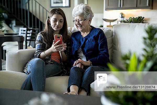 Enkelin zeigt der Großmutter ein Smartphone  während sie zu Hause auf dem Sofa sitzt