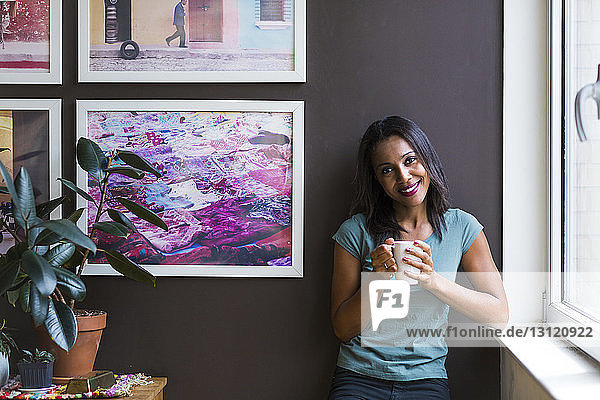 Porträt einer Frau  die eine Kaffeetasse in der Hand hält  während sie zu Hause vor einem Bilderrahmen an der Wand steht