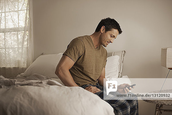 Lächelnder Mann telefoniert  während er zu Hause auf dem Bett sitzt