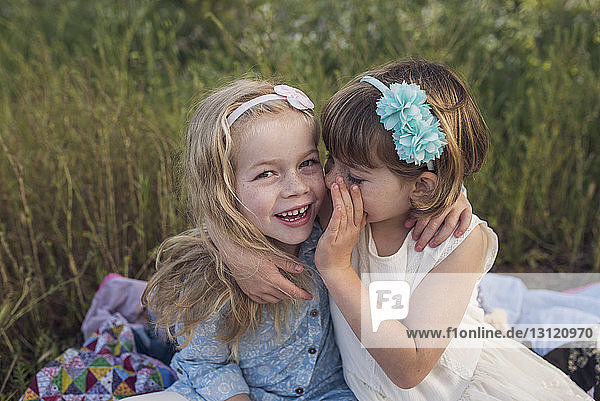 Hochwinkelaufnahme eines Mädchens  das der Schwester ins Ohr flüstert  während sie im Park auf einer Decke sitzt