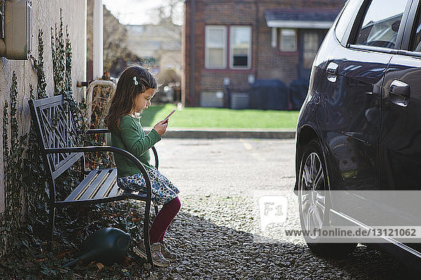 Side view of girl using mobile phone while sitting on bench at sidewalk by car