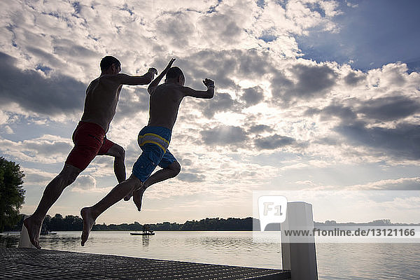 Father and son jumping from pier at lake against sky