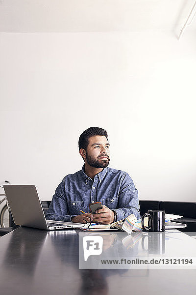 Thoughtful man with laptop and mobile phone sitting at table against wall