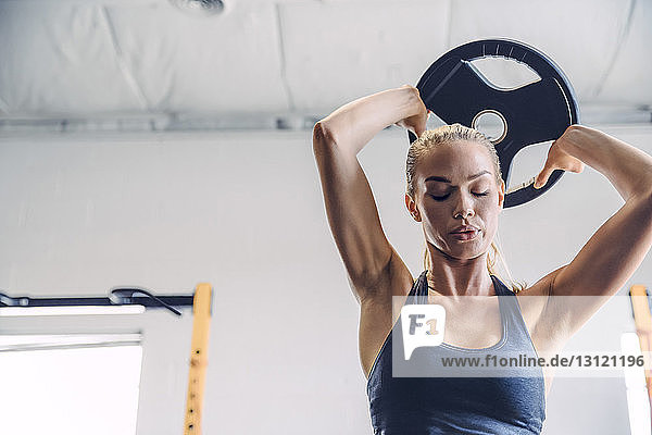 Woman with eyes closed lifting weight plate while exercising in gym