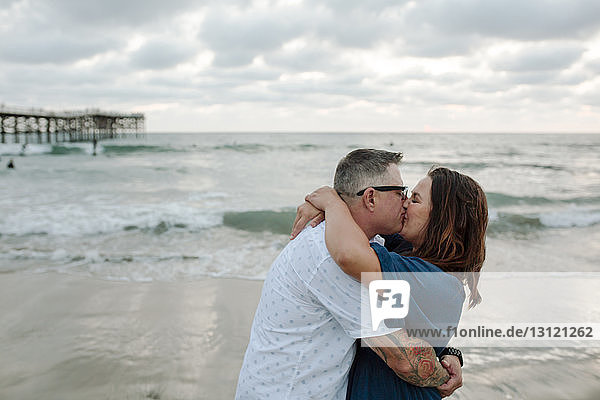 Seitenansicht eines Paares  das sich am Strand vor bewölktem Himmel küsst