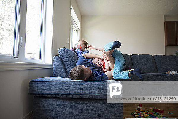 Glücklicher Vater spielt mit dem Sohn  während die Tochter zu Hause auf dem Sofa Wasser trinkt