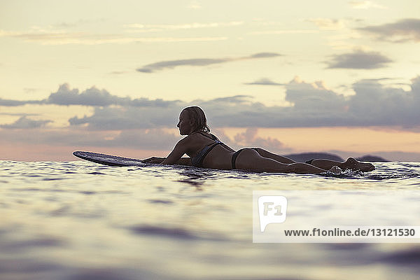 Seitenansicht einer sinnlichen Frau  die bei Sonnenuntergang auf einem Surfbrett im Meer gegen den Himmel liegt