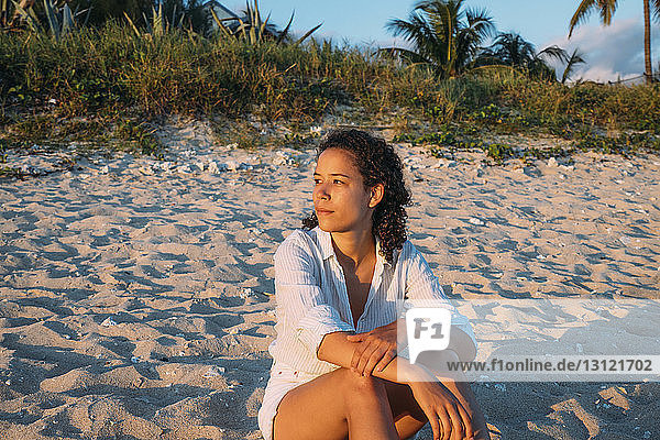 Frau schaut weg  während sie bei Sonnenuntergang am Strand auf Sand sitzt