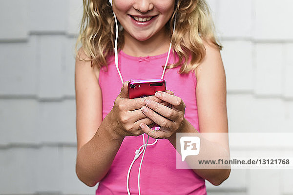 Midsection of happy girl listening music while using smart phone against wall