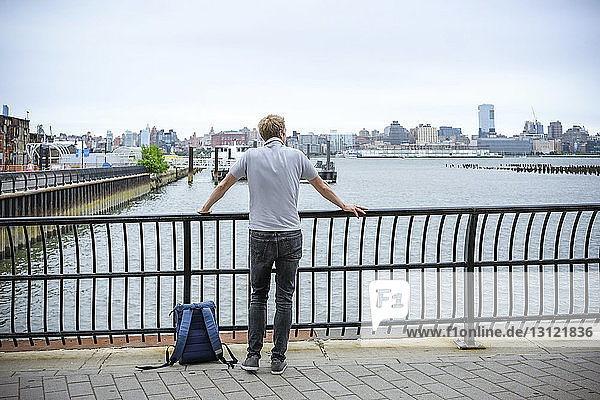 Rear view of man with backpack looking at river while standing on promenade against sky in city