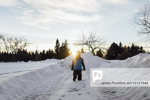 Rear view of boy walking on snow covered field against sky during sunset