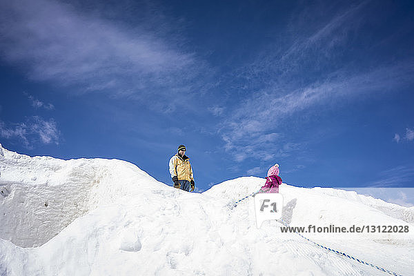 Niedrigwinkelaufnahme eines Vaters mit Tochter auf einem schneebedeckten Hügel vor blauem Himmel während eines sonnigen Tages