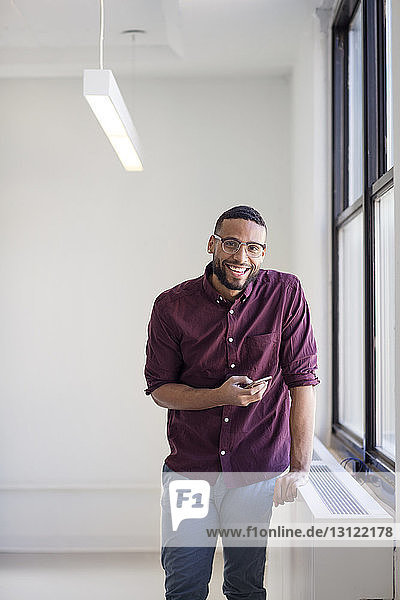 Porträt eines Geschäftsmannes mit Mobiltelefon in der Hand  während er in einem neuen Büro am Fenster steht