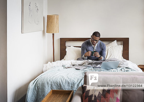 man holding coffee cup while reading newspaper on bed at home