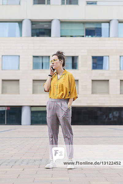 Selbstbewusste Geschäftsfrau spricht mit einem Smartphone  während sie auf dem Bürgersteig gegen ein Gebäude in der Stadt steht