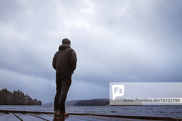 Man looking at river while standing on jetty against cloudy sky