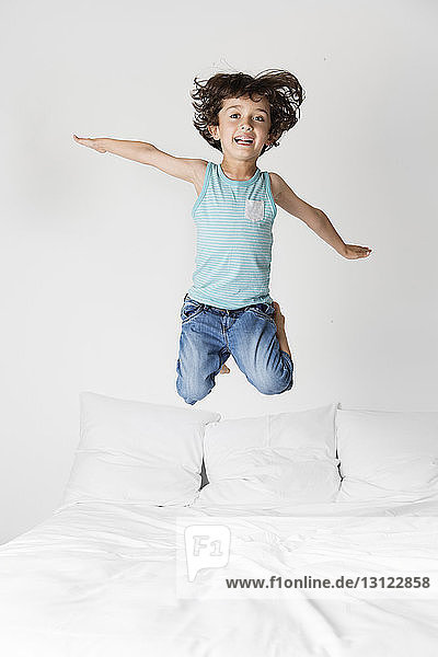 Portrait of happy boy with arms outstretched jumping on bed against wall at home